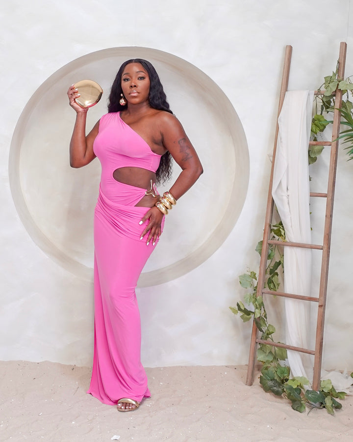 Woman in a pink one-shoulder dress holding a gold bowl in a minimalistic setting with a ladder and plant.